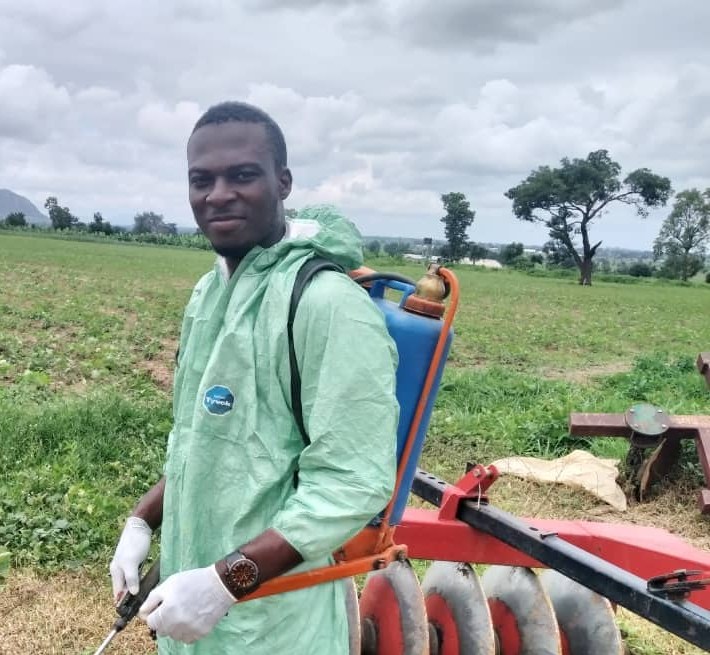 Emmy in protective coveralls with a backpack sprayer beside a disc harrow